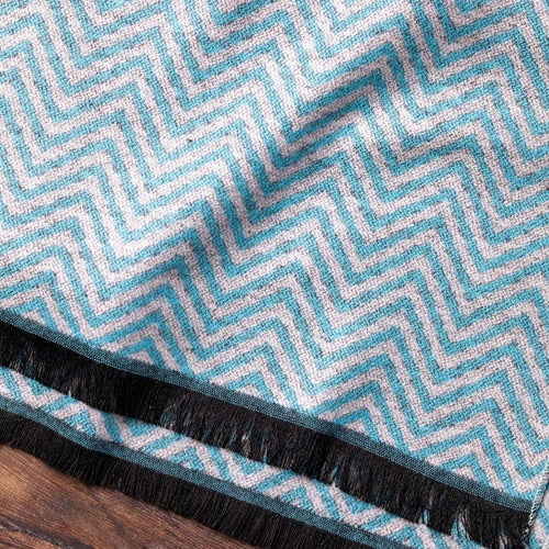 Close-up of a blue and white zigzag patterned fabric with black fringe on a wooden surface.