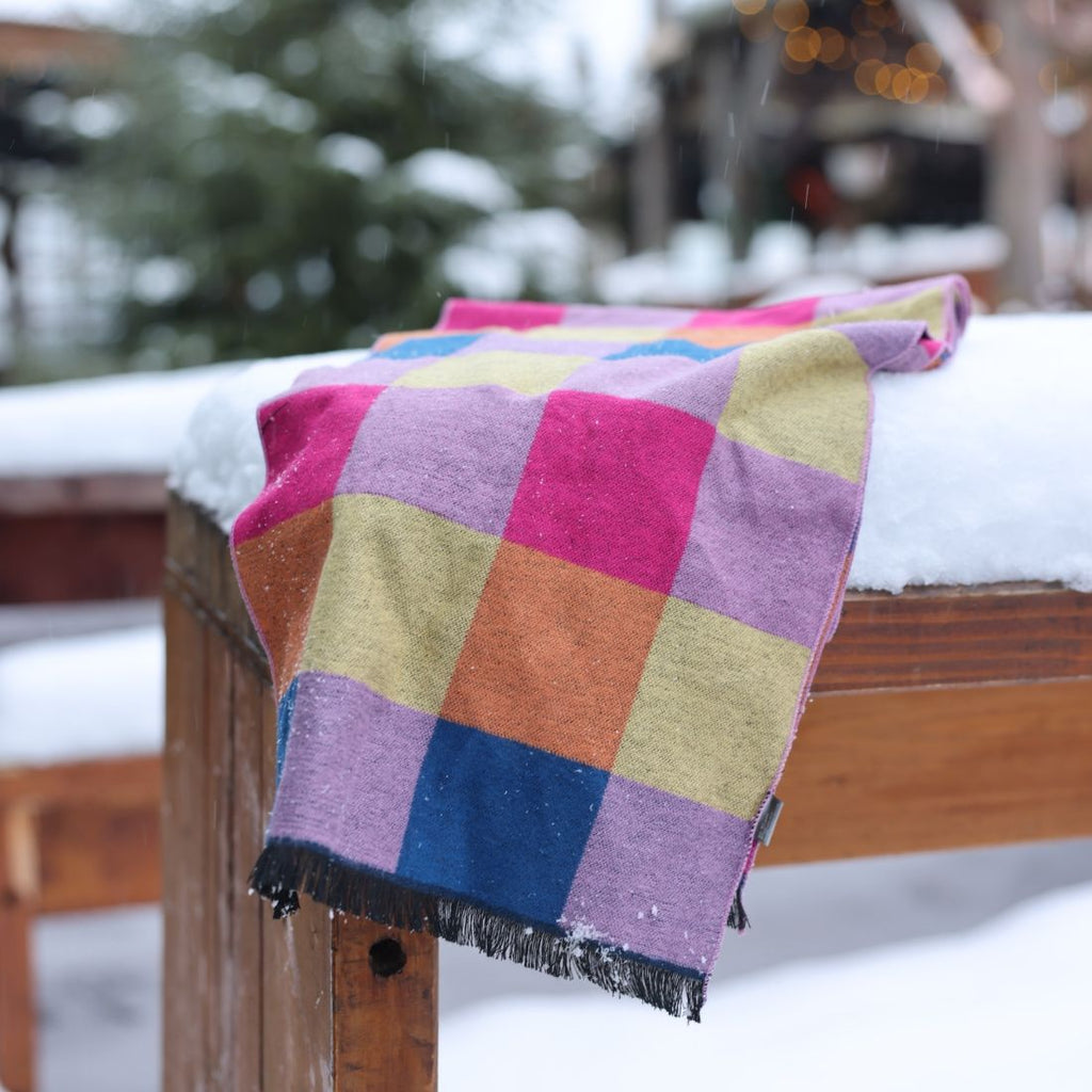 Colorful checkered blanket draped over a wooden bench in a snowy outdoor setting.