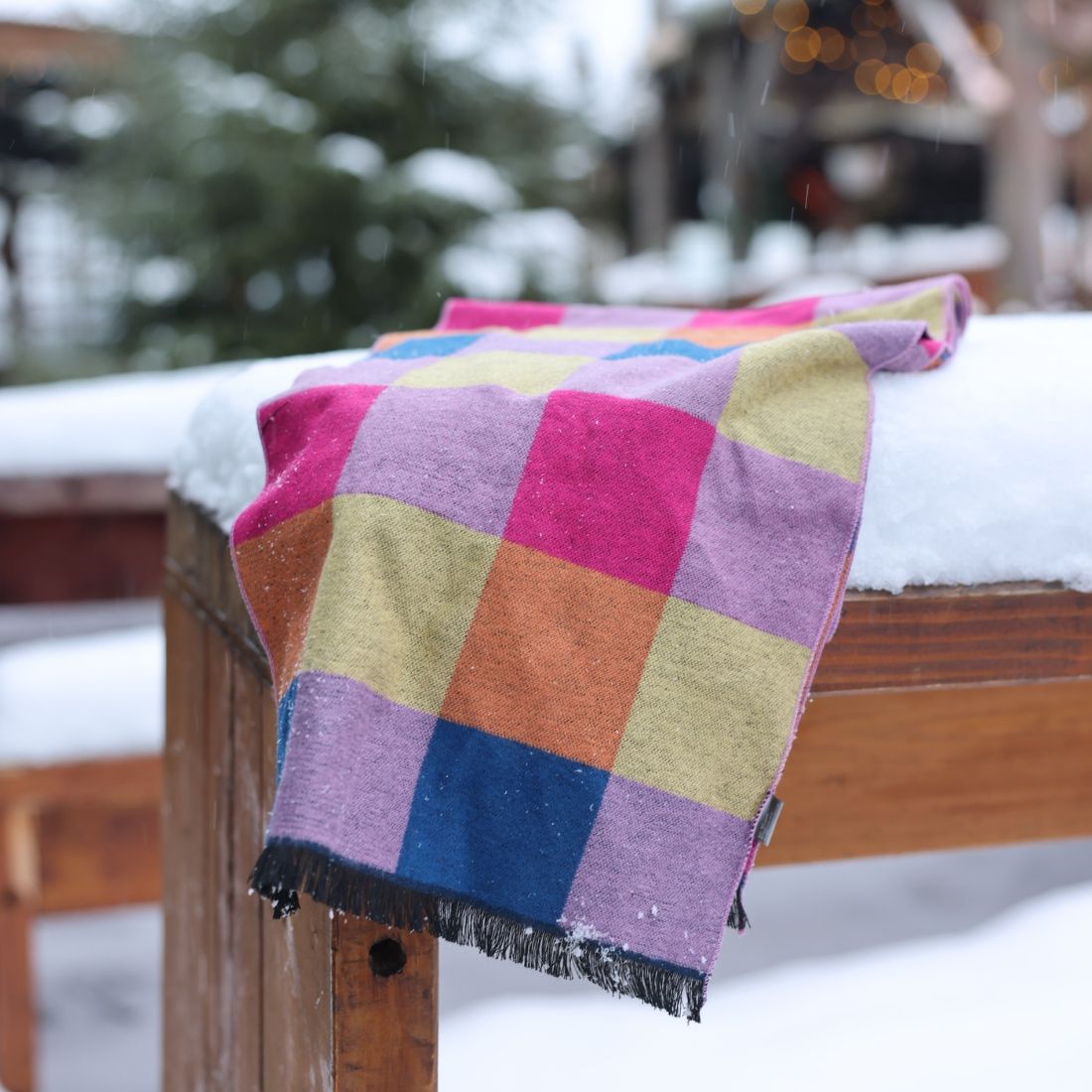 Colorful checkered blanket draped over a wooden bench in a snowy outdoor setting.