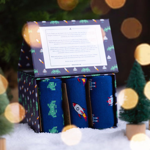 Box of socks with space-themed designs in front of a Christmas tree with lights.