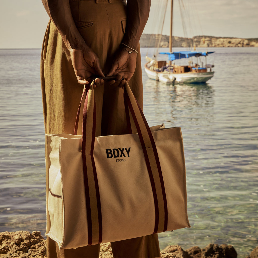 Person holding a beige bag with 'BDXY' branding by the water with a sailboat in the background.