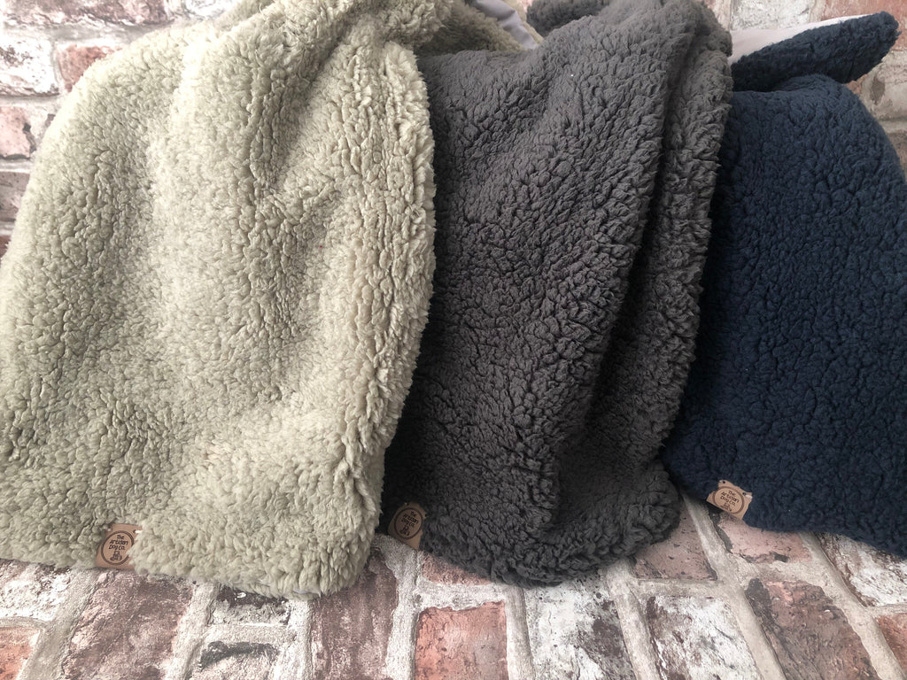 Three folded fleece blankets in green, gray, and black on a brick surface.