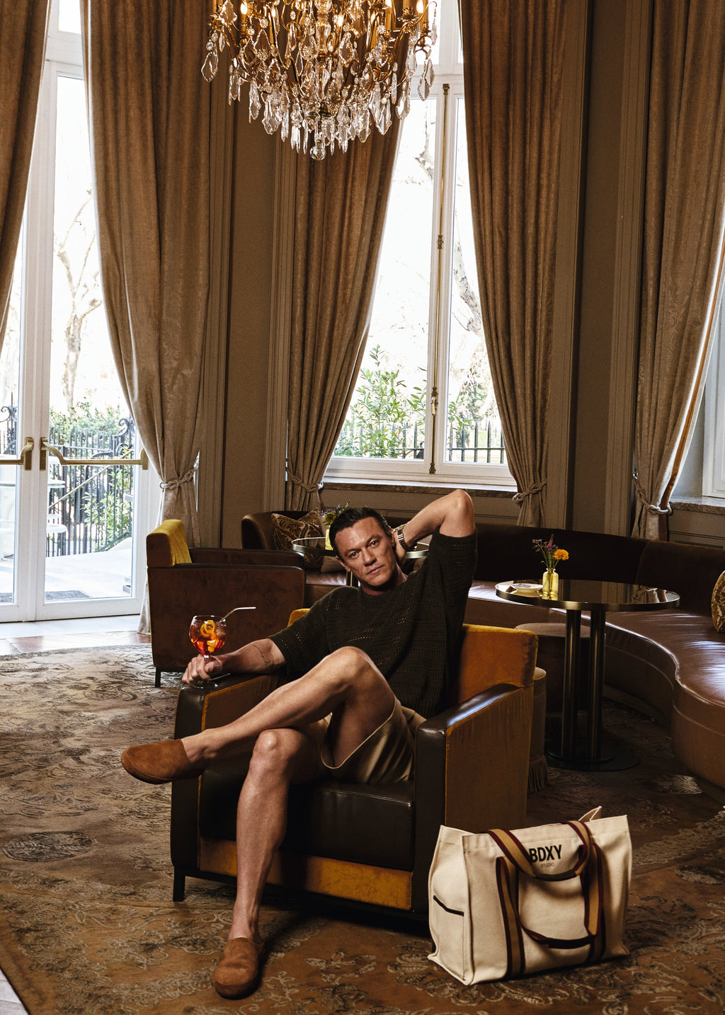Man sitting in a luxurious room with chandelier and large windows, next to a 'Goyard' bag.