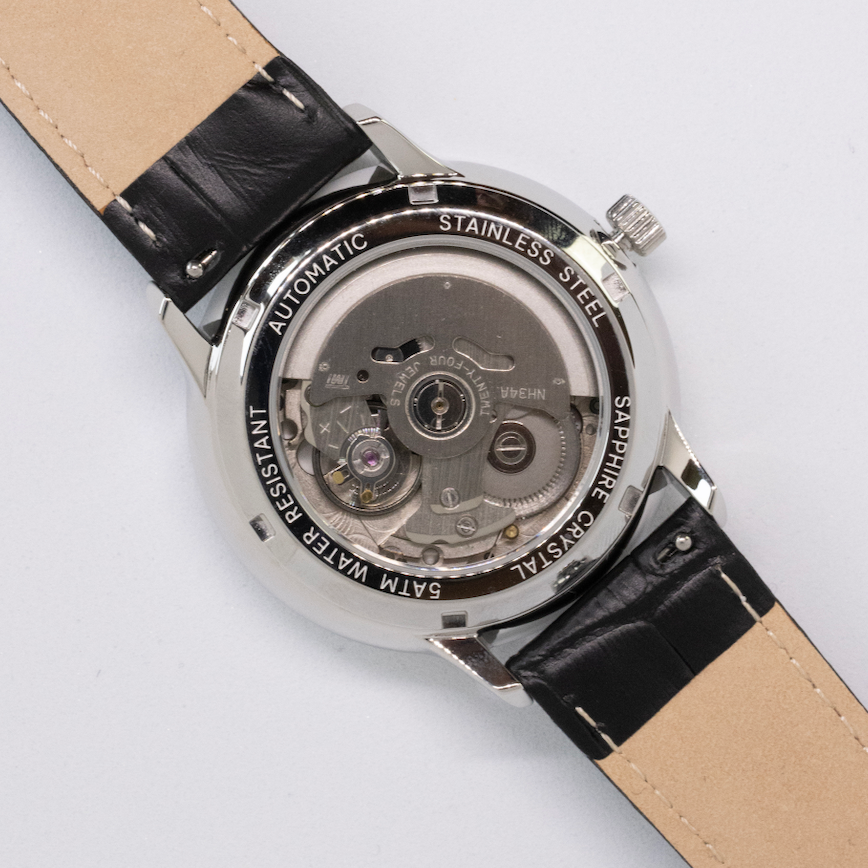 Close-up of a watch with a black strap and visible mechanical components on a light gray background