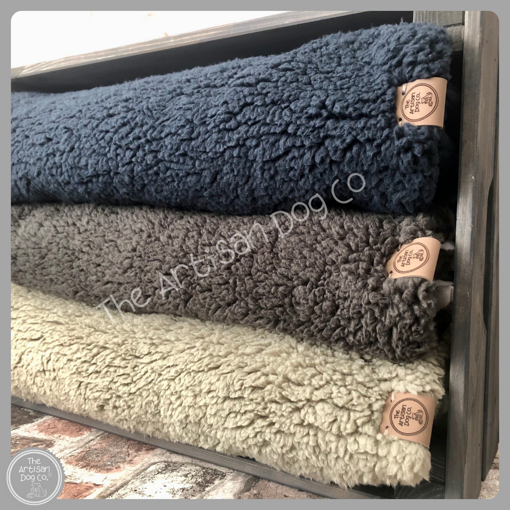 Stack of blue, gray, and beige dog blankets with 'The Artisan Dog Co' labels.