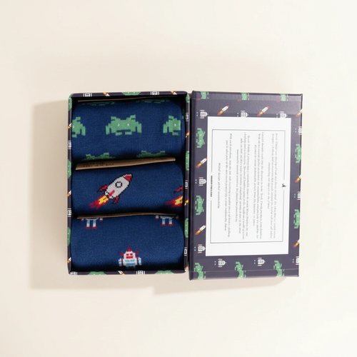 Three pairs of socks with space-themed designs in an open box on a light beige background